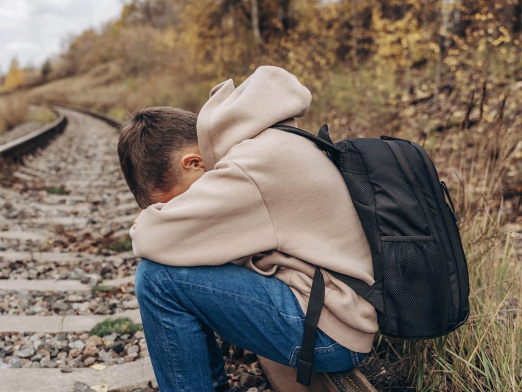 A distressed-looking boy sits on train tracks in the countryside. He wears a tan hoodie, bluejeans, and a black backpack. He has his head resting on his arms, crossed over his knees while he sits.