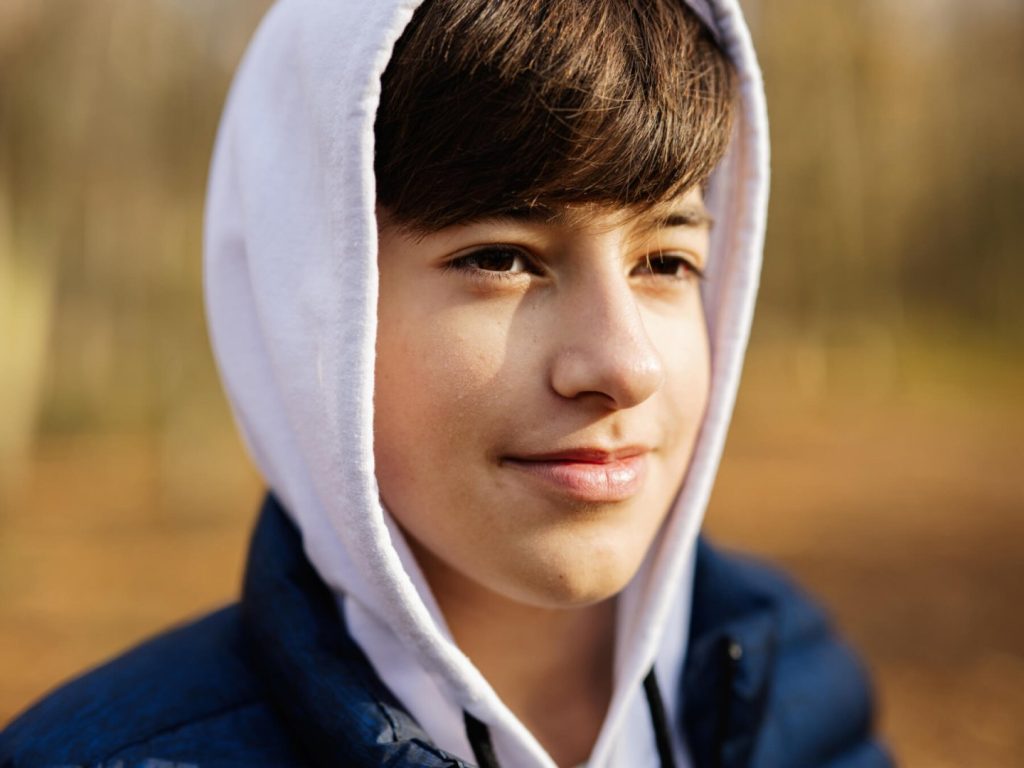 A close-up portrait of a boy outside. He has brown hair and eyes and smiles slightly as he looks off in front of him.