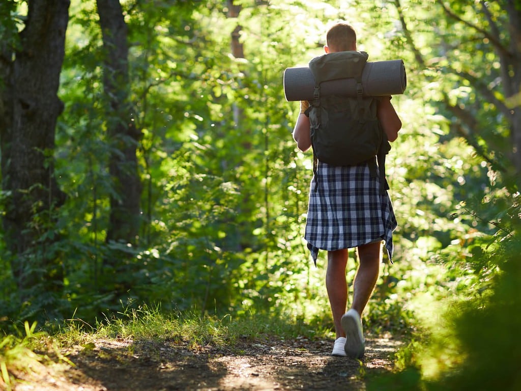 A man with a striped flannel tied around his waist carries a camping pack and bed roll down forest trail at golden hour.
