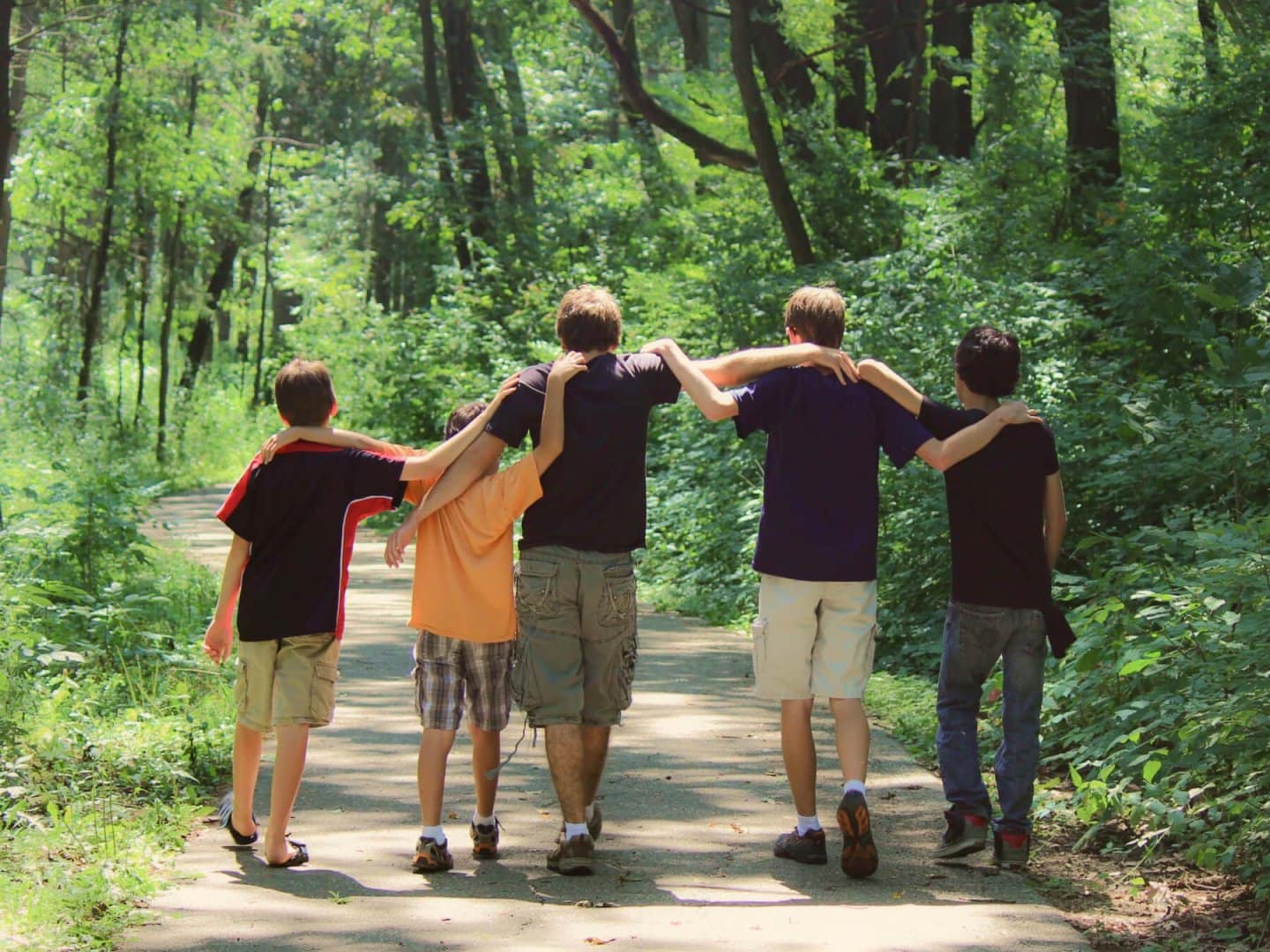 Five boys of varying height, age, and race walk with their arms over each others shoulders down a trail in a forest.