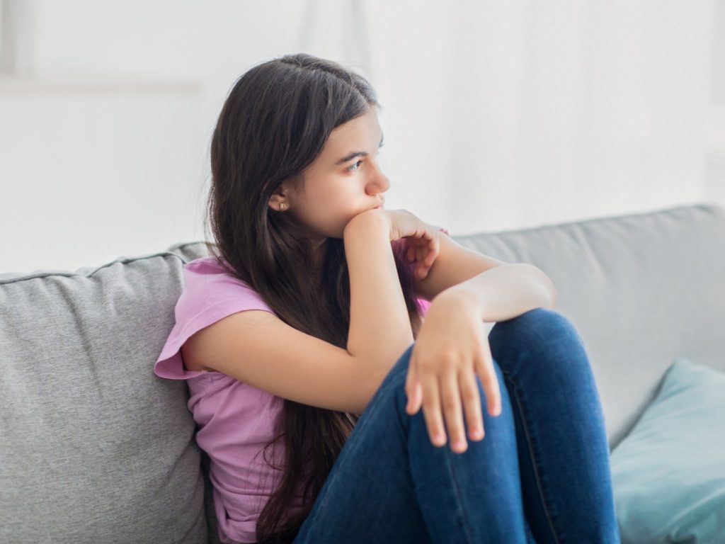 A teen girl with long brown hair sits with her knees up and arms crossed on a grey sofa, looking away from the viewer.