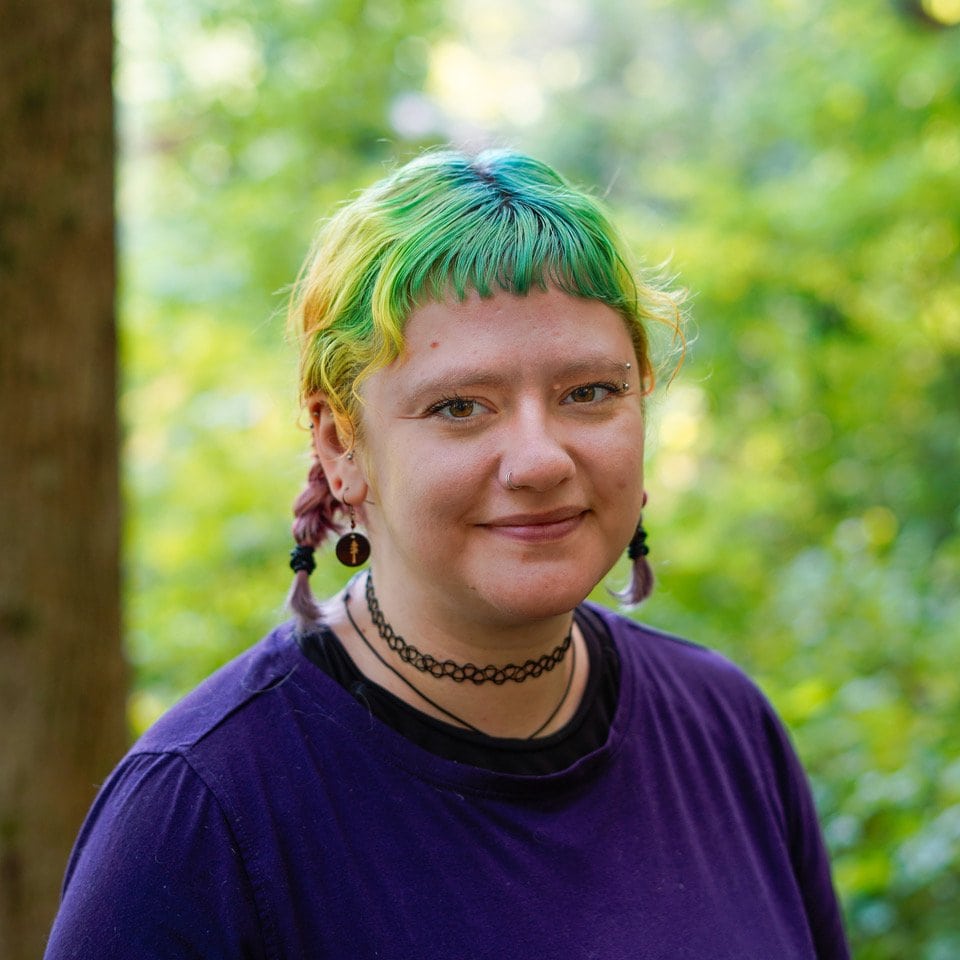 Julia Ruff, a Primary Therapist at Trails Carolina, wears a purple shirt and rainbow-dyed hair in two braids and smiles at the camera.