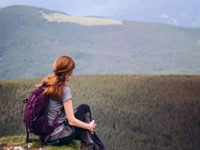 A red-headed young woman sits on a hilltop overlooking forested mountains in front of her.