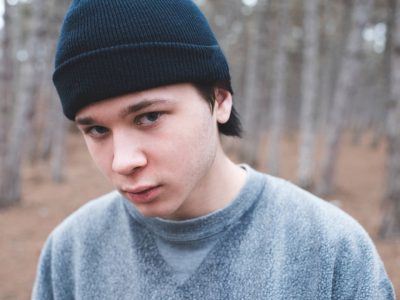 A teen boy in a beanie and a sweatshirt glowers at the viewer in the woods.