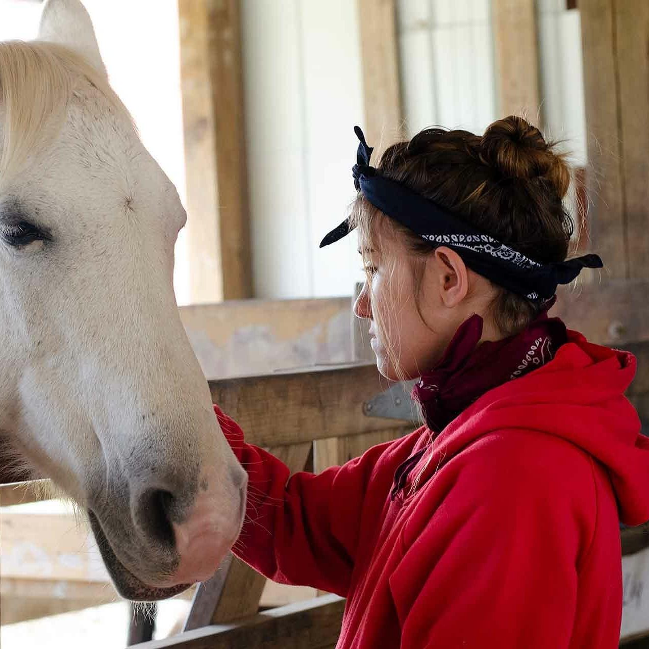 Youth behavioral camps -adolescent girl wearing a red coat and bandana headband pets a white horse.